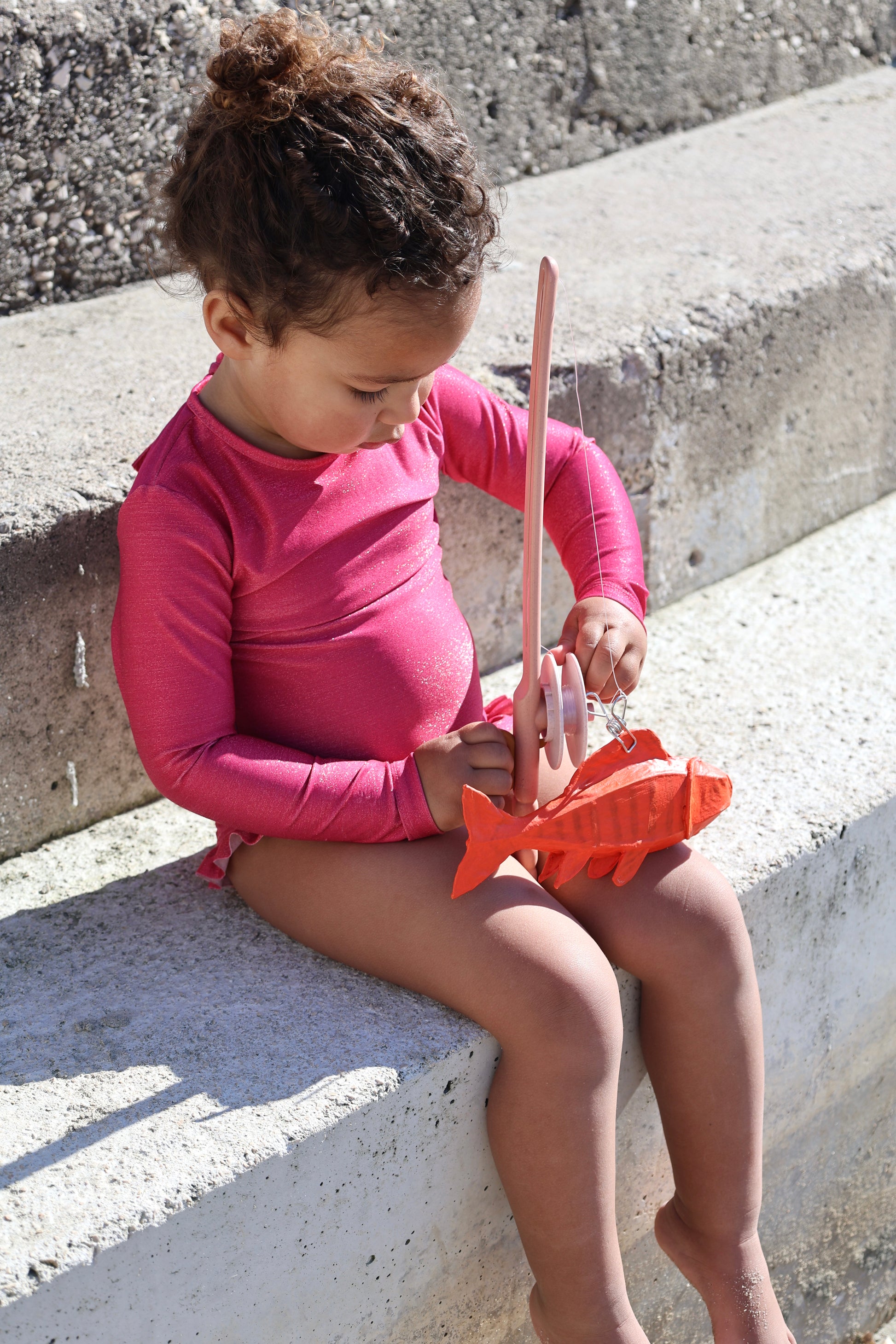 Baby girl wearing Loumio Paris Alba raspberry swimsuit with fish details, playing on the beach